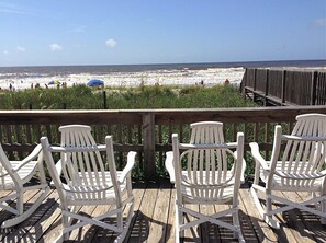 A wooden deck with white Adirondack chairs overlooking a beach with people and umbrellas. - Beautiful OIB Condo- 1 Min. Walk to Beach; includes Pools, Tennis Courts, Extras (Ocean Isle Beach)