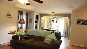 Un salon chaleureux avec un canapé marron, un ventilateur de plafond et une cuisine visible au fond.