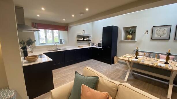 A modern kitchen with dark cabinetry, a central island, and a dining area with a table set for four. - Rectory Farm Barn (Grantham)