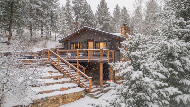A cabin with a wooden staircase leading to a deck, surrounded by snow-covered trees.