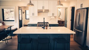 A modern kitchen with white cabinets, a central island with bar stools, stainless steel appliances, pendant lighting, and a view of the outdoors.