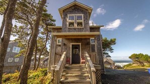 Une maison en bois avec un porche à l’avant et des marches menant à la porte, entourée d’arbres et offrant une vue sur l’océan.