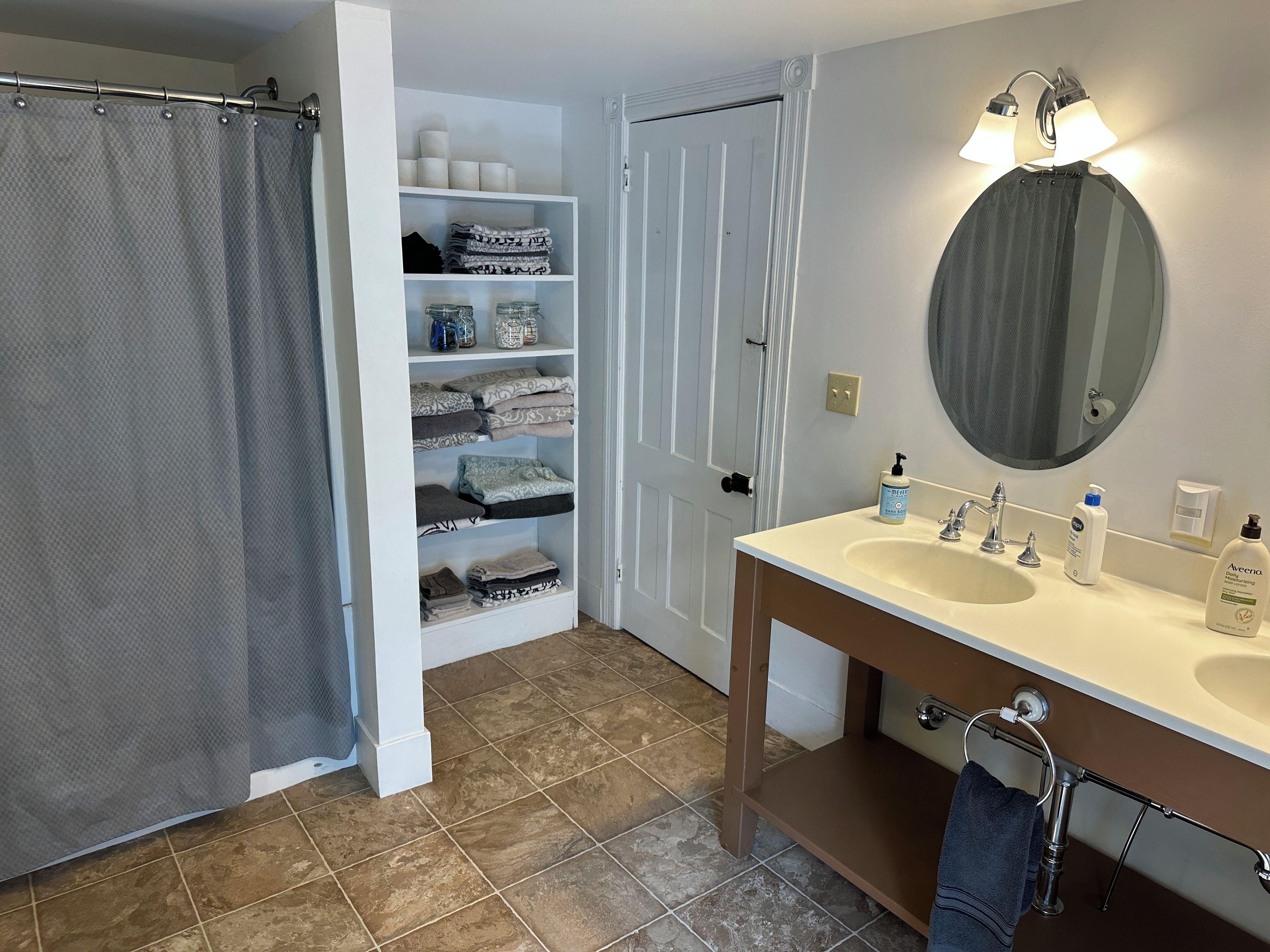 A bathroom with a shower curtain, a shelf with folded towels, a vanity with a mirror, and toiletries.