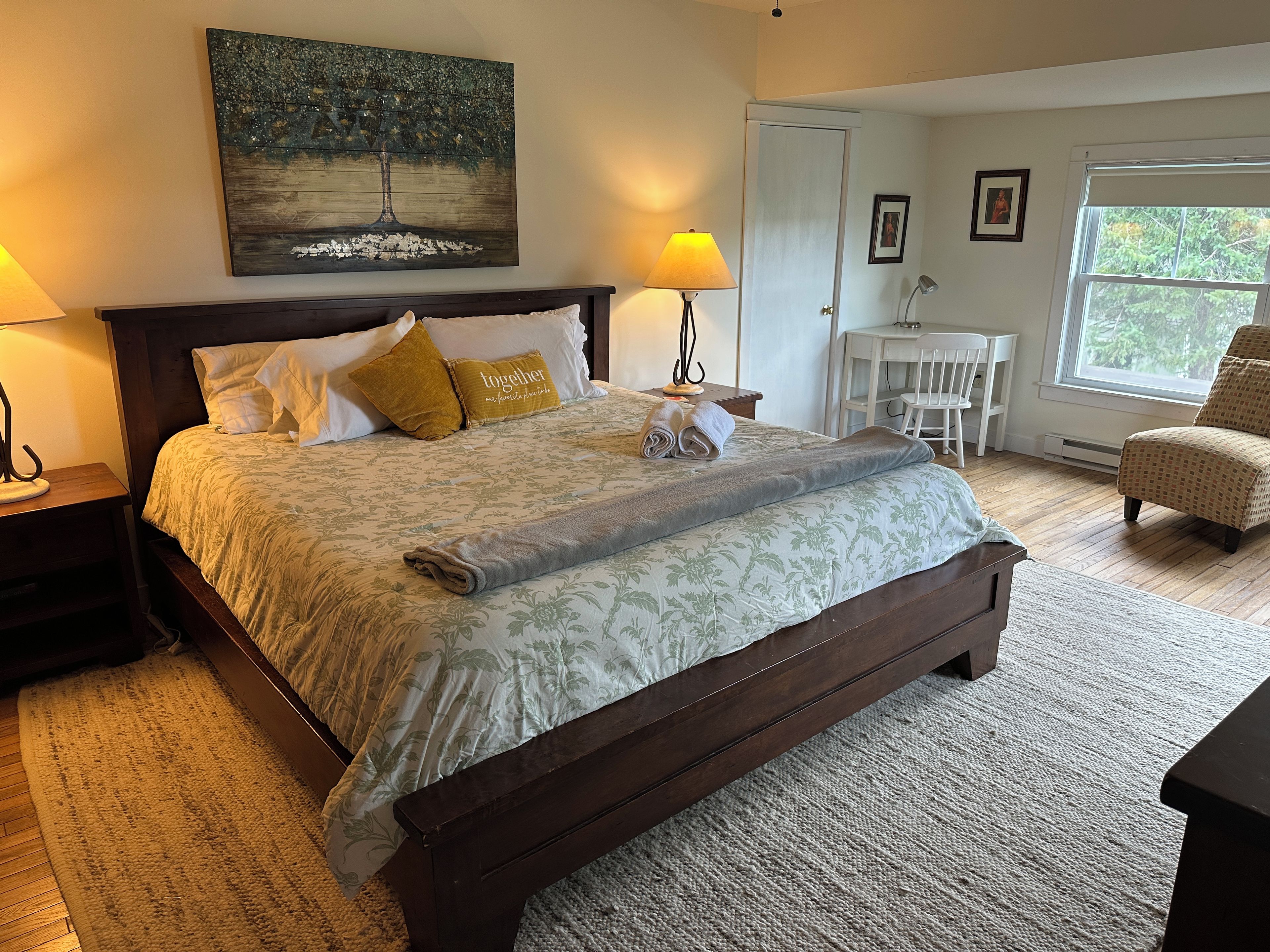 A bedroom with a large bed, a nightstand, a desk, a chair, a window, and a painting on the wall.