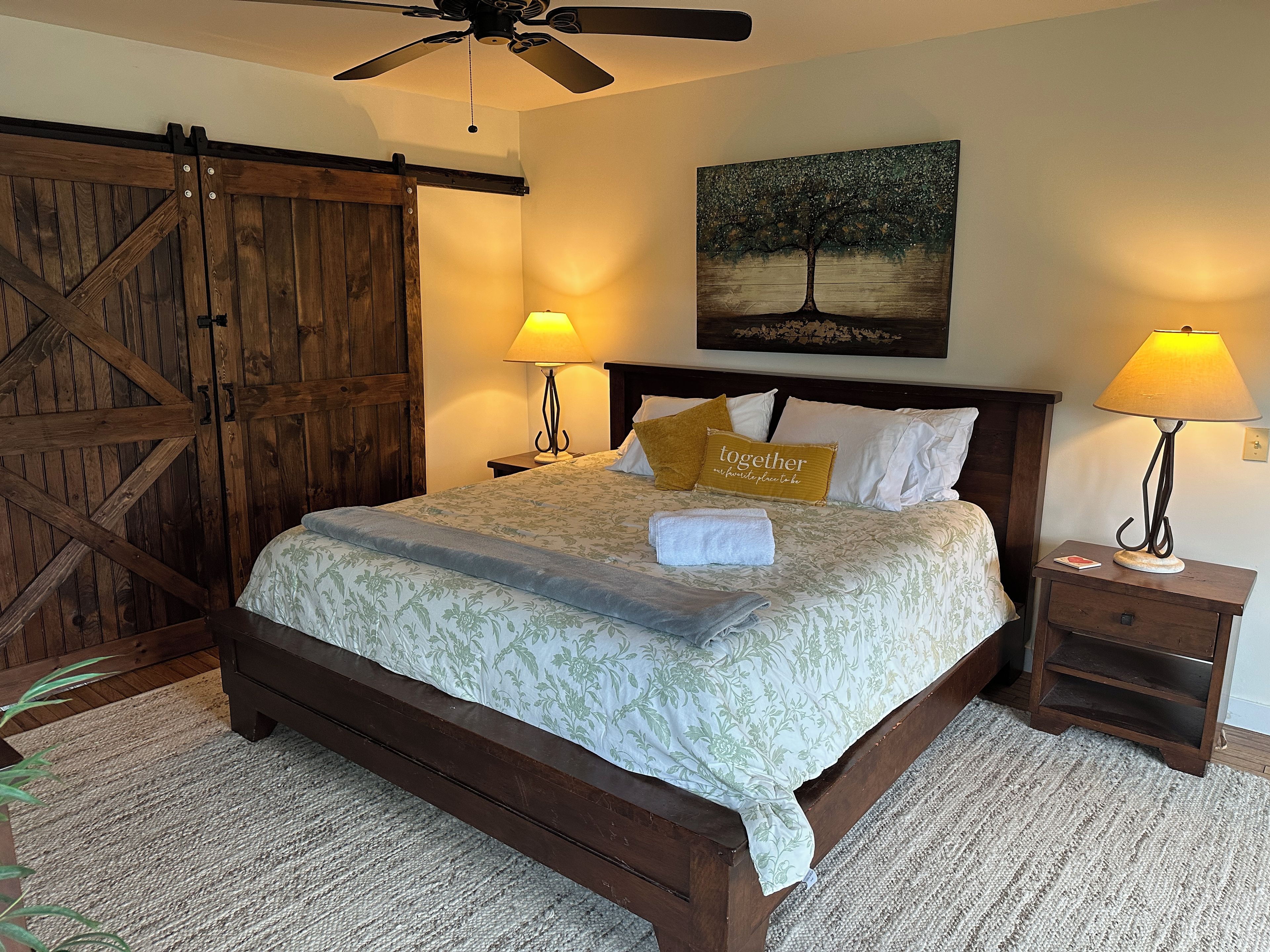 A bedroom with a wooden bed, two bedside lamps, a painting on the wall, and a ceiling fan.