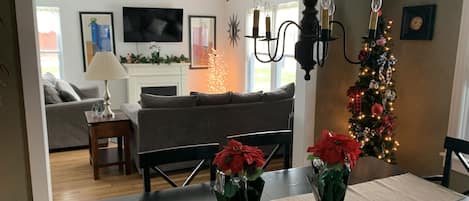 Un comedor con una mesa dispuesta para dos, un árbol de Navidad y una sala de estar al fondo.