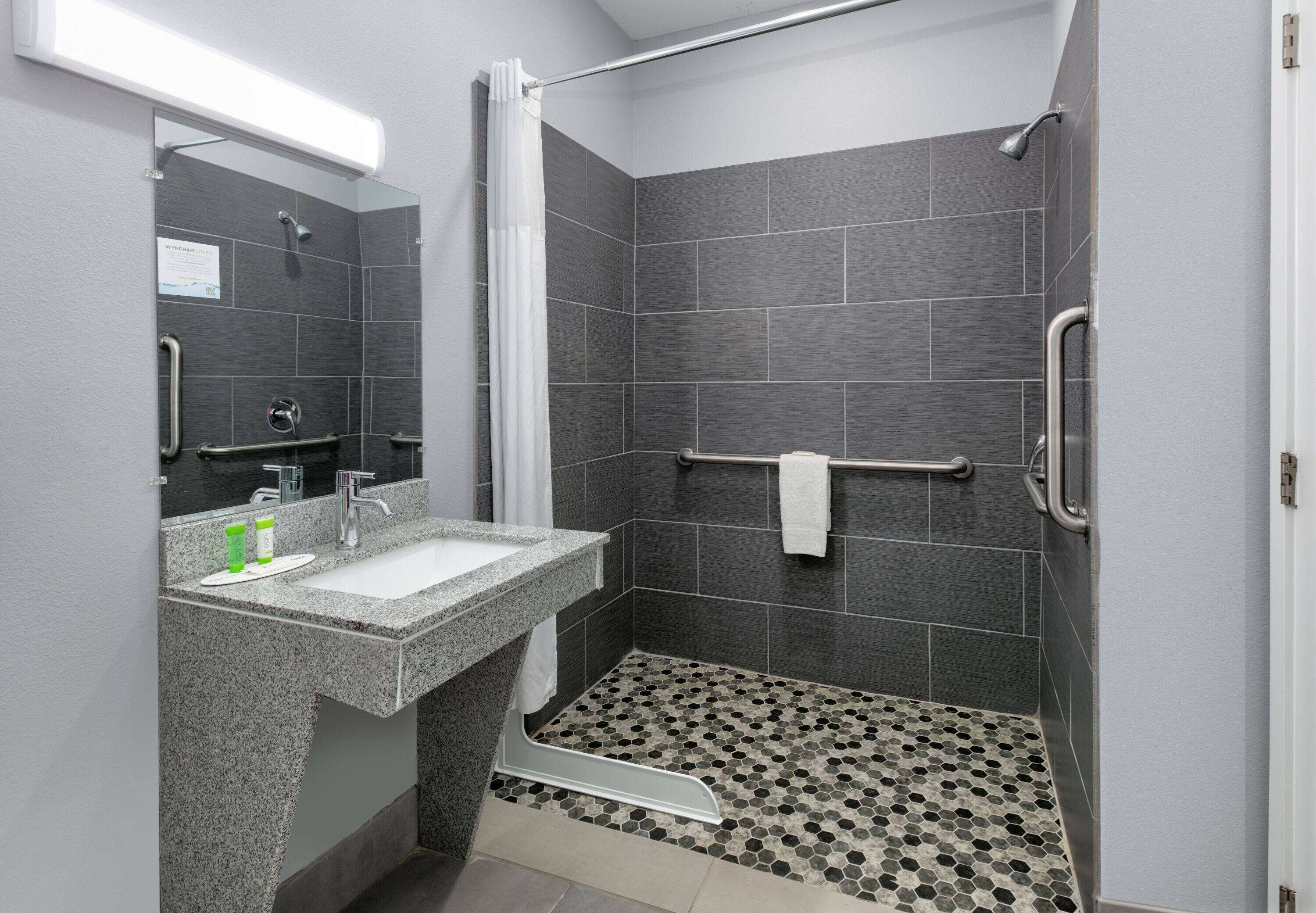Een moderne badkamer met een granieten wastafel, een spiegel, een douche met een handdouche en een betegelde vloer met een honingraatpatroon.