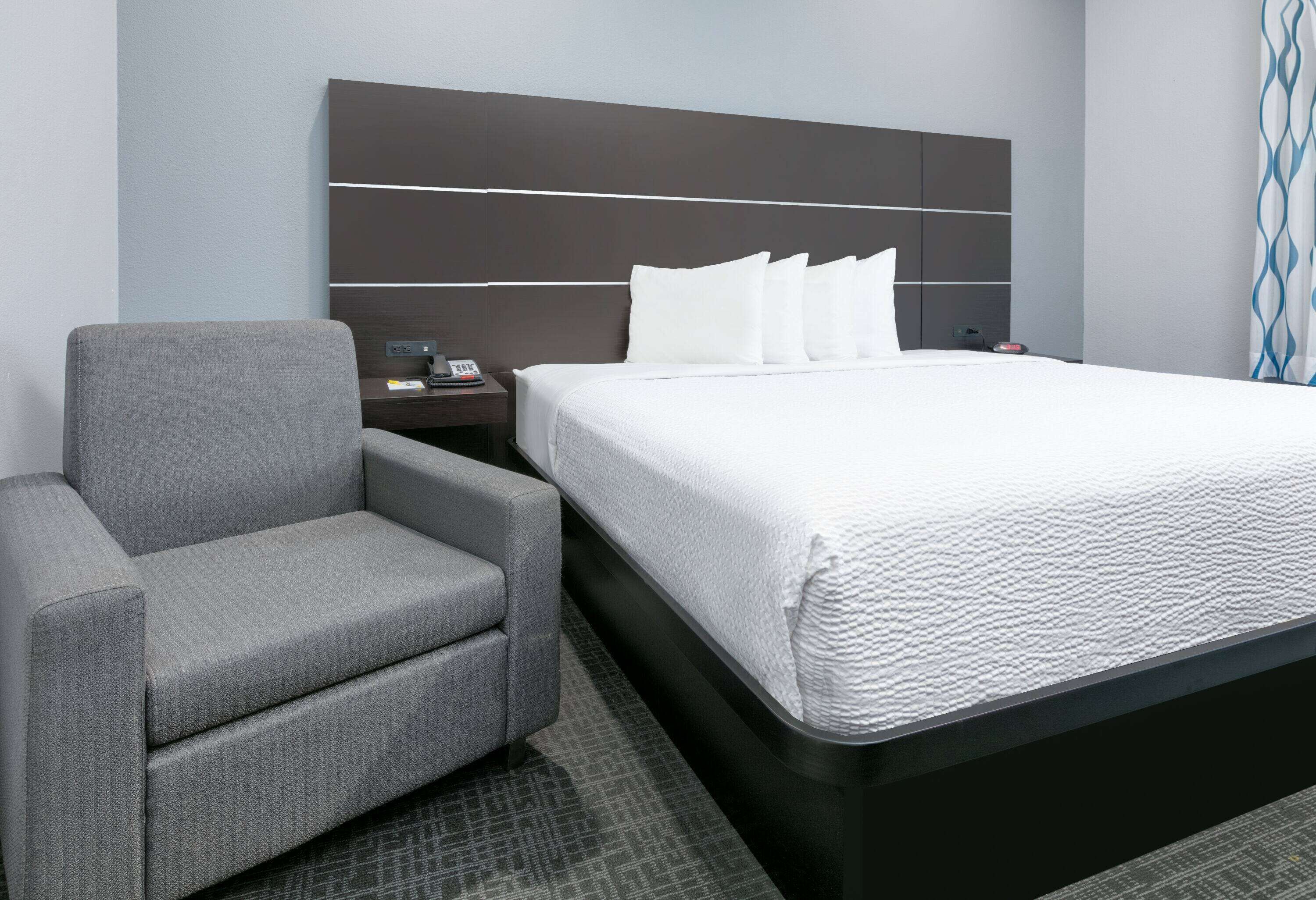 Ein Hotelzimmer mit einem Bett, einem grauen Sessel, einem Nachttisch mit Telefon und Lampe sowie einem modernen Kopfteil.