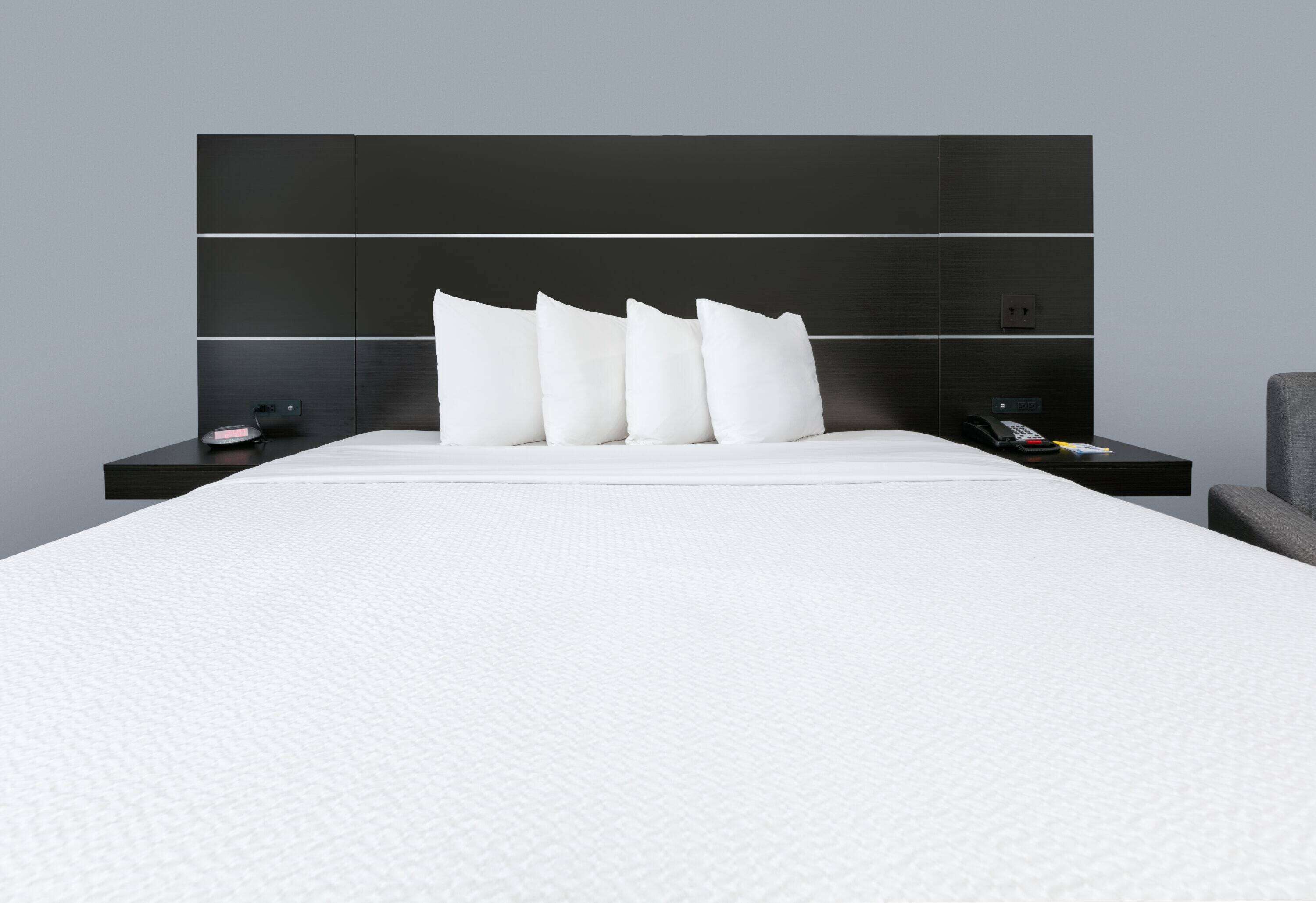 A modern hotel room with a large bed, a headboard with horizontal lines, and a bedside table with a phone and a small lamp.