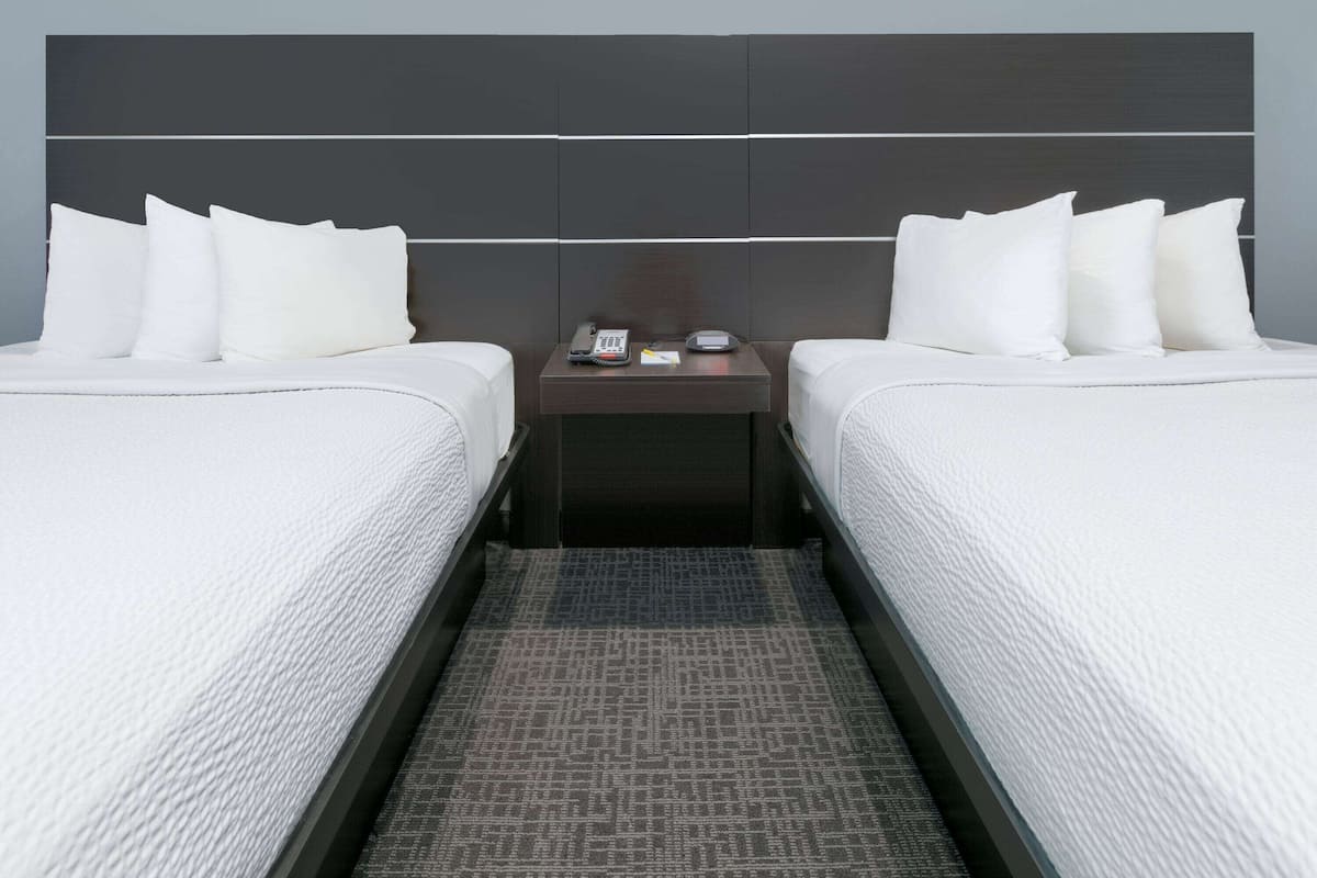 A hotel room with two beds, a nightstand with a phone and a clock, and a patterned rug.