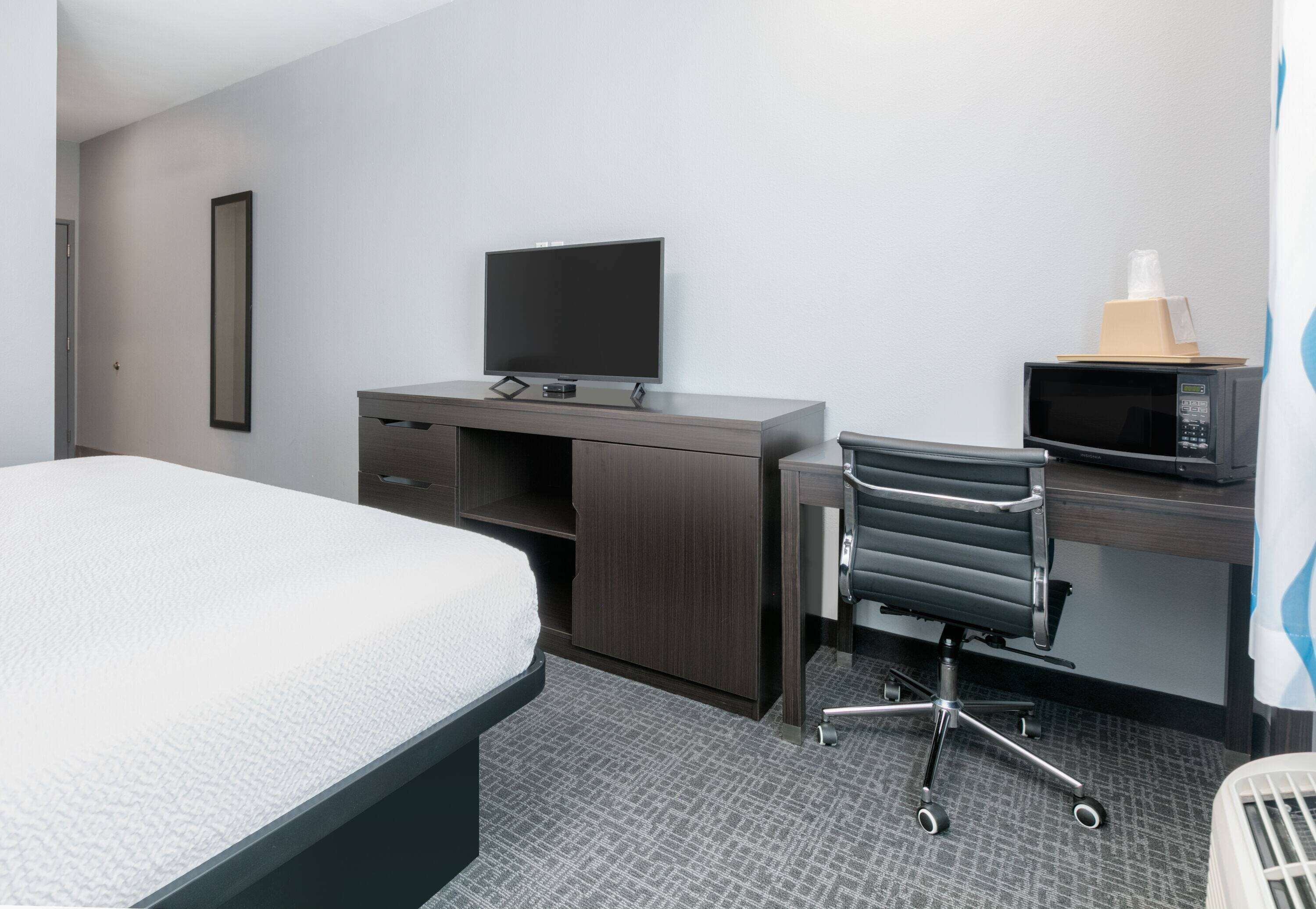 A hotel room with a bed, a desk with a flat-screen TV, a chair, a microwave, and a small table with a tissue box.