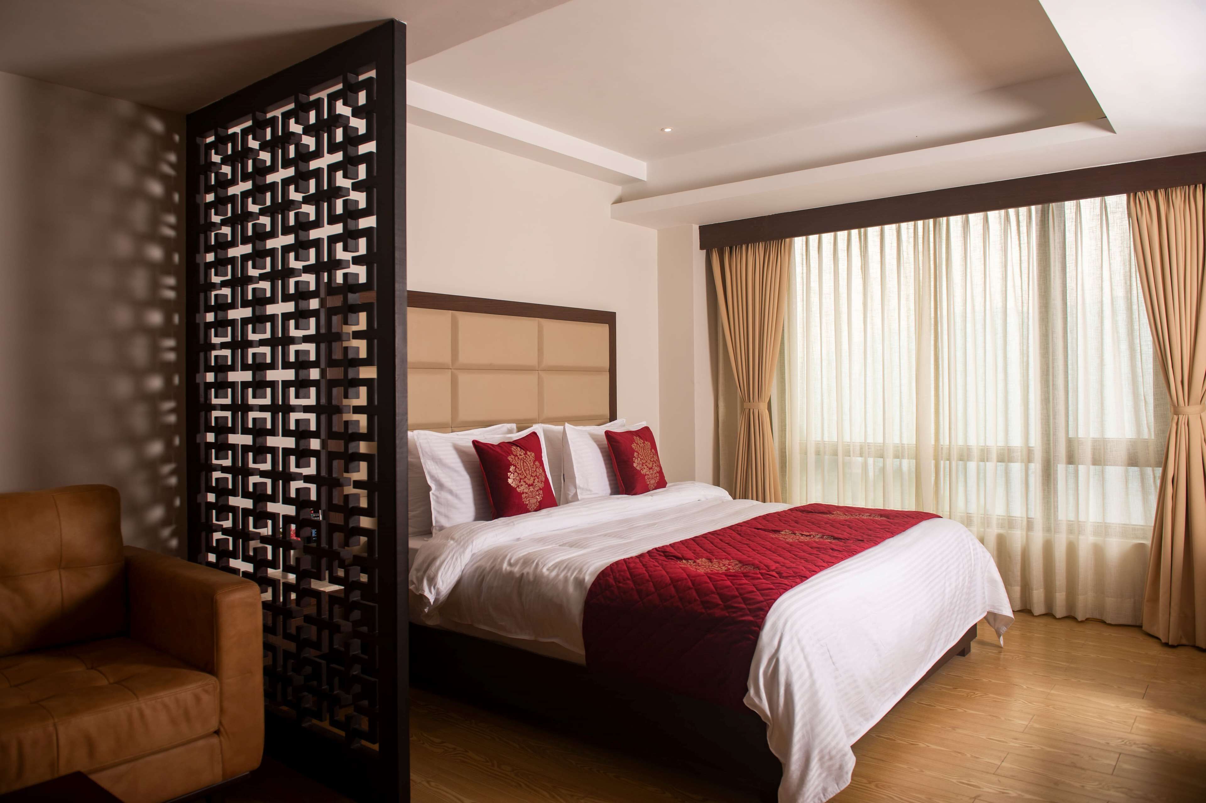 Una habitació d'hotel amb un llit, un sòl de fusta, un separador decoratiu i una finestra amb cortines.