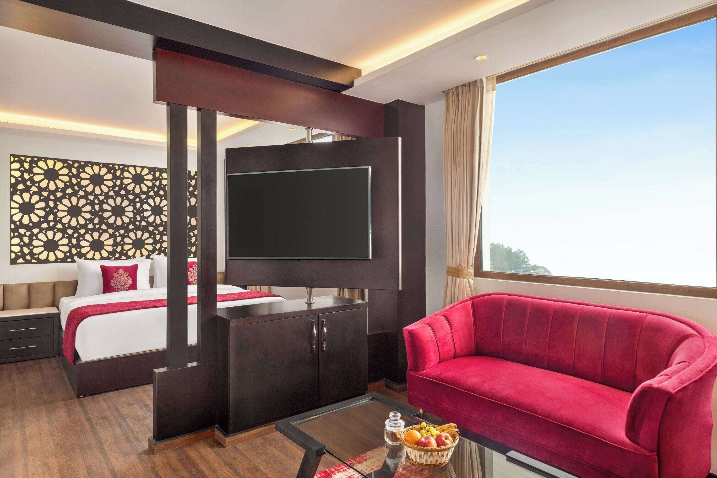 A hotel room with a bed, a TV, a red sofa, and a large window.