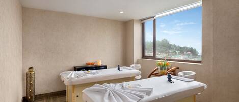 Un cuarto de spa con dos camillas, un ventanal con vistas a la ciudad y un jarrón decorativo con flores.
