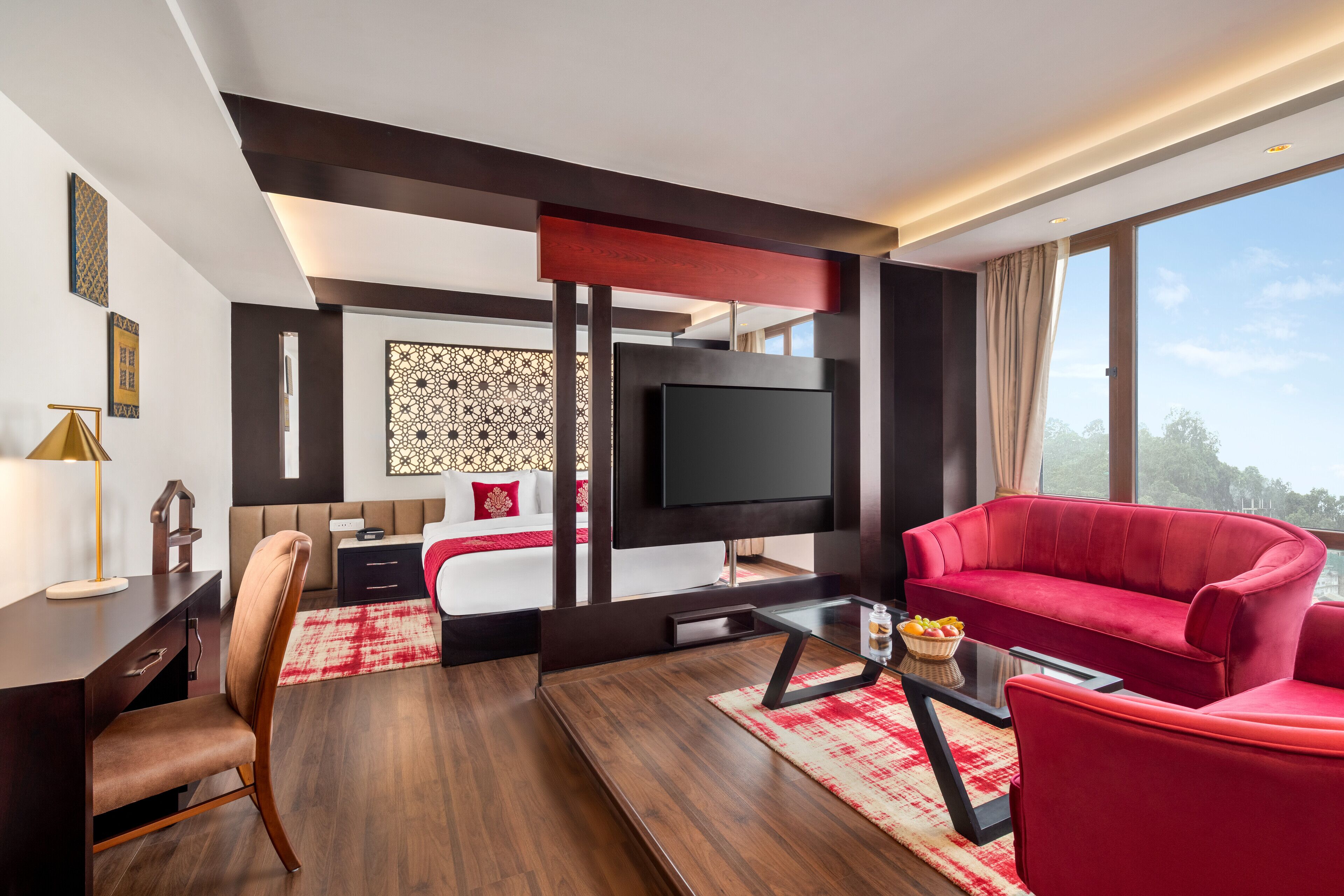 A modern hotel room with a large TV, a red sofa, a wooden desk, and a large window offering a city view.