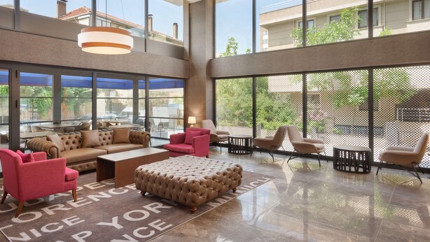 Un vestíbulo amplio con techo alto, ventanas grandes y una zona de estar con otomano capitonado y sillas modernas.