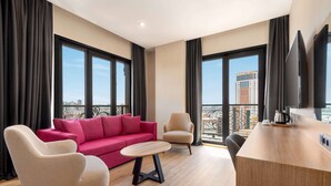 Una habitación de hotel moderna con un sofá rojo, dos sillones, una mesa de centro de madera, televisión y ventanas amplias con vistas a la ciudad.