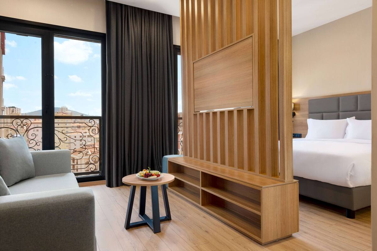 A hotel room with a sofa, a small round table with a fruit bowl, a TV mounted on a wooden panel, a bed with white bedding, and a view of buildings through large windows.