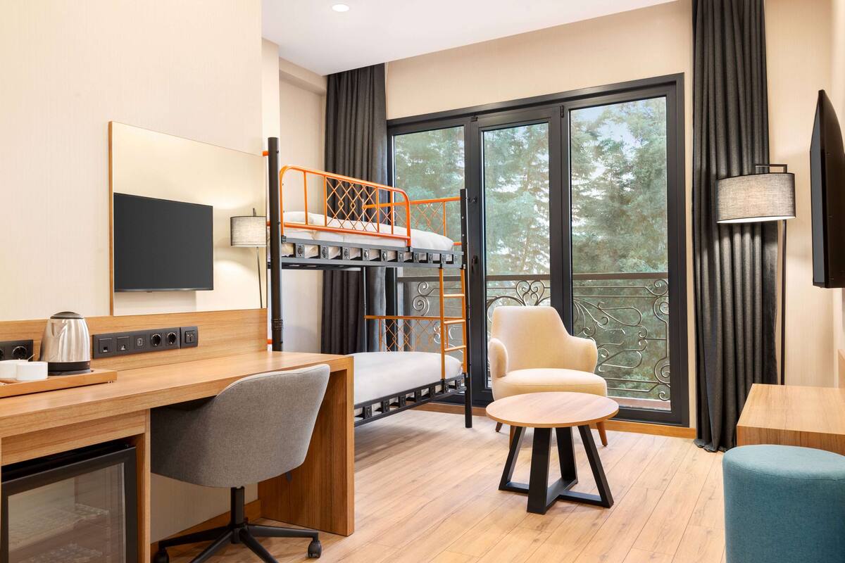 A hotel room with a bunk bed, a desk with a chair, a small table, a TV, and a window with curtains.
