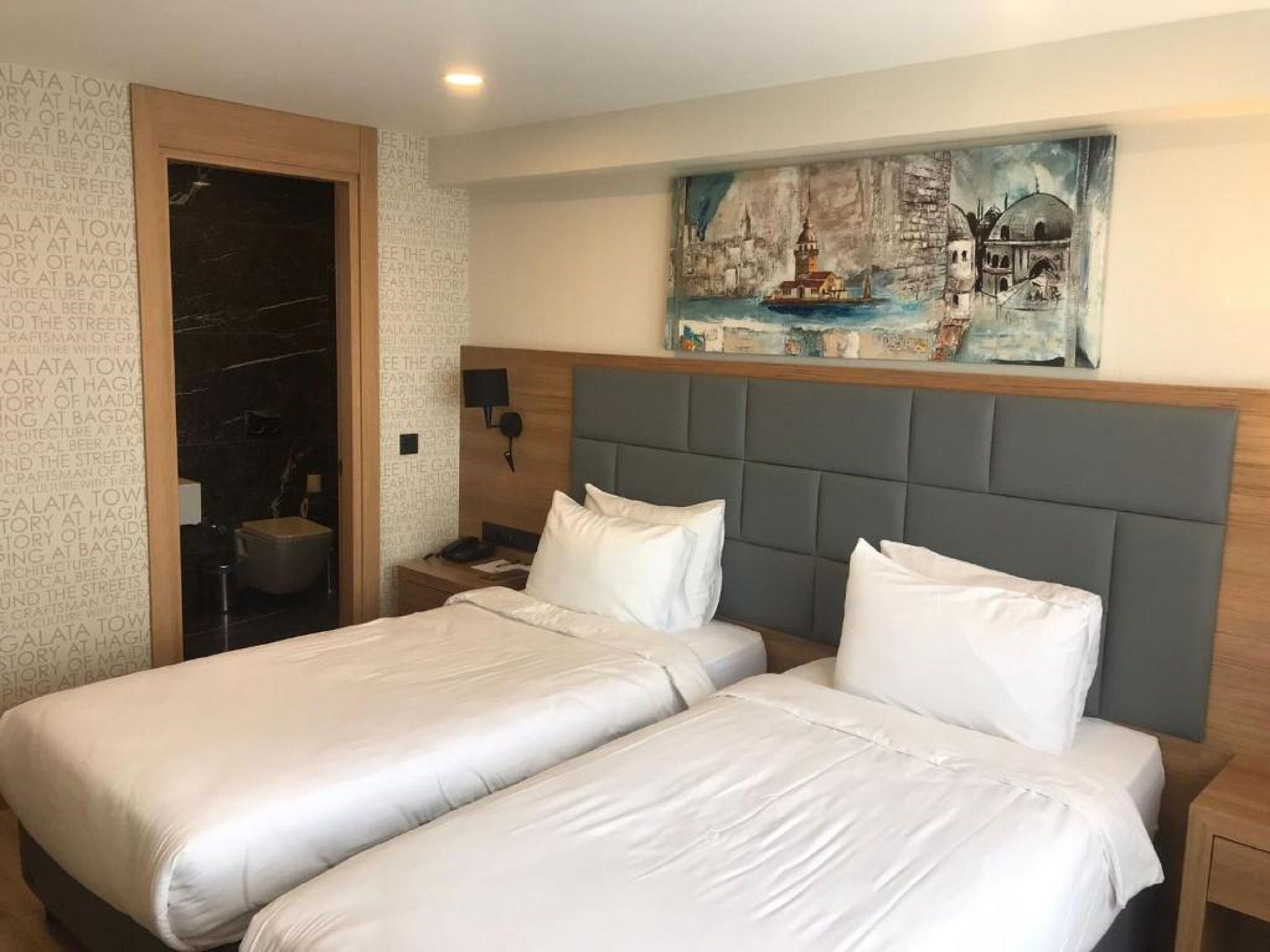 A hotel room with two beds, a headboard, a painting of a coastal scene, and a wall with text.