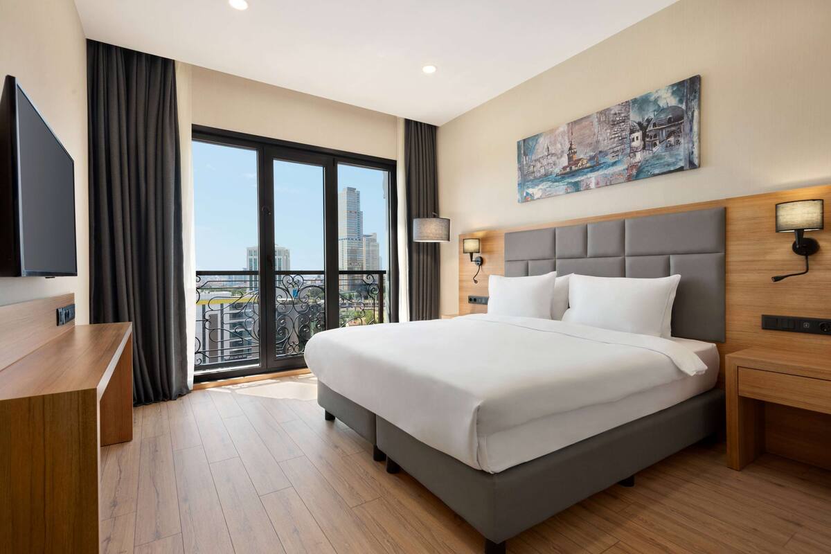 A modern hotel room with a large bed, a desk, and a balcony with a city view.