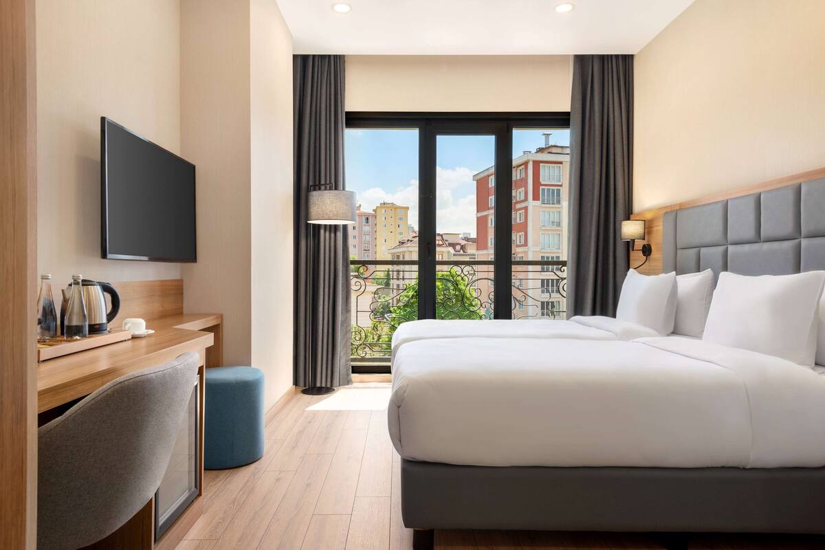 A hotel room with a bed, a desk with a chair, a TV, and a balcony with a view of buildings.