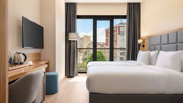 A hotel room with a bed, a desk with a chair, a TV, and a balcony with a view of buildings.