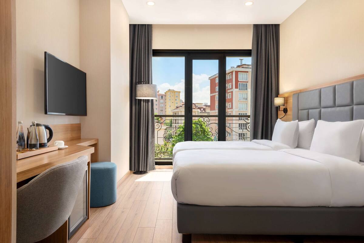 A hotel room with a bed, a desk, a chair, a TV, and a balcony with a view of buildings.