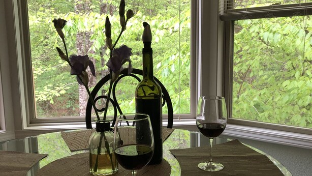 A dining table with a glass top, surrounded by woven placemats, a wine bottle, and a glass of red wine.