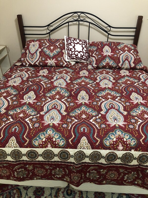 Una cama con colcha estampada y almohadas, cabecera de metal y pared blanca.