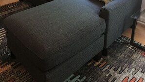 A living room with a dark grey sectional sofa, two patterned throw pillows, a floor lamp, and a window with floral curtains.