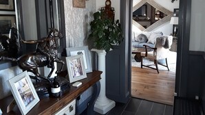 Une pièce avec une table en bois sur laquelle sont disposées des photos encadrées et une bouteille de vin, ainsi qu’une grande plante en pot et un escalier menant à une autre pièce.