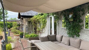 Un espace extérieur aménagé avec une longue table et des bancs rembourrés, entouré de plantes en pot et doté d’un patio couvert.