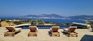 Área à beira da piscina com espreguiçadeiras e vista para o mar e montanhas.