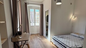 Ein Zimmer mit Holzboden, einem Bett mit Matratze und Kissen, einem Schreibtisch mit Stuhl, einer Hängeleuchte und einer Tür zu einem anderen Raum.