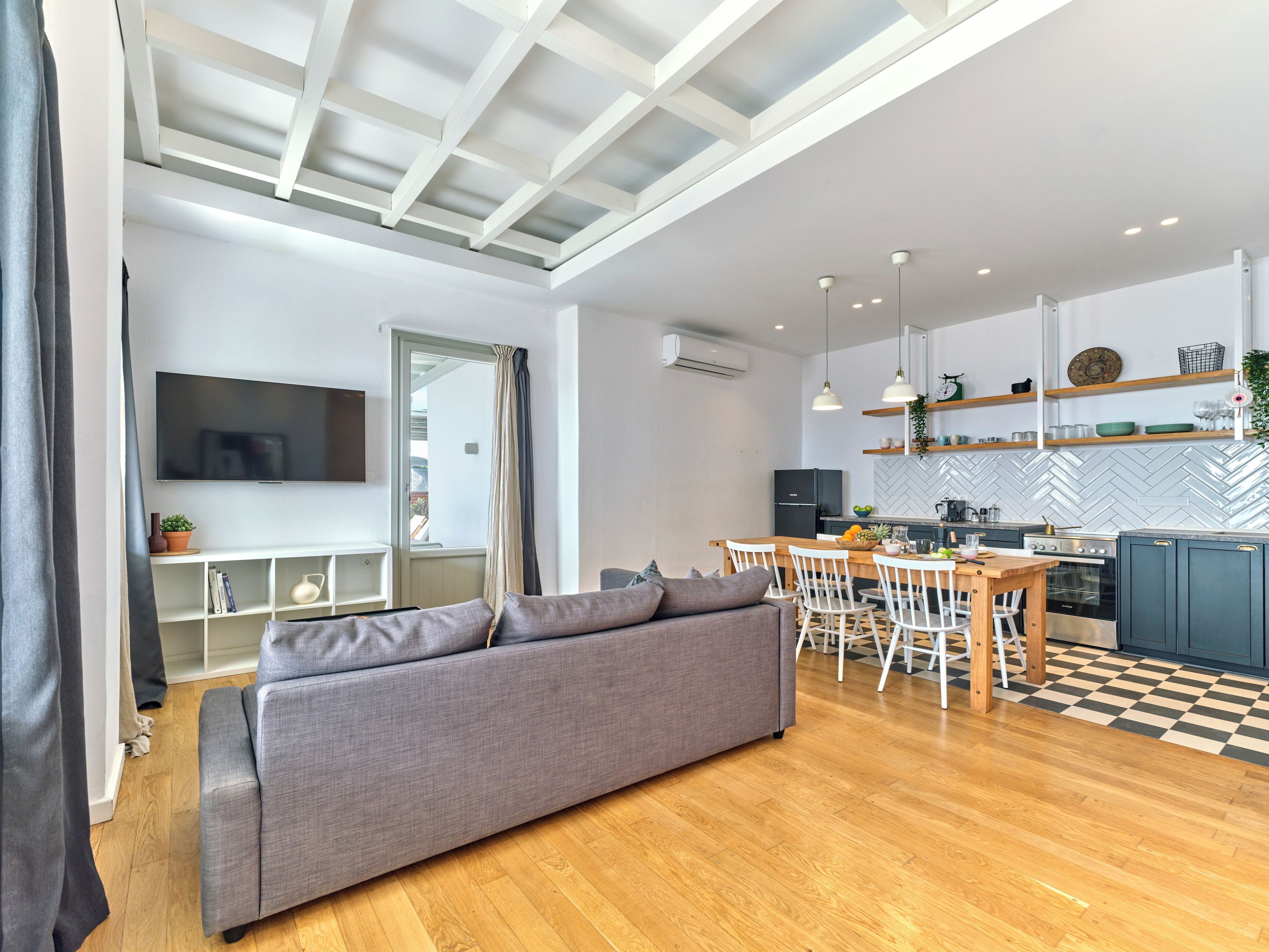 Un salon moderne avec un canapé gris, une table à manger en bois et des chaises. Un téléviseur est fixé au mur, et il y a un coin cuisine avec un dosseret de crédence à motifs en chevrons.