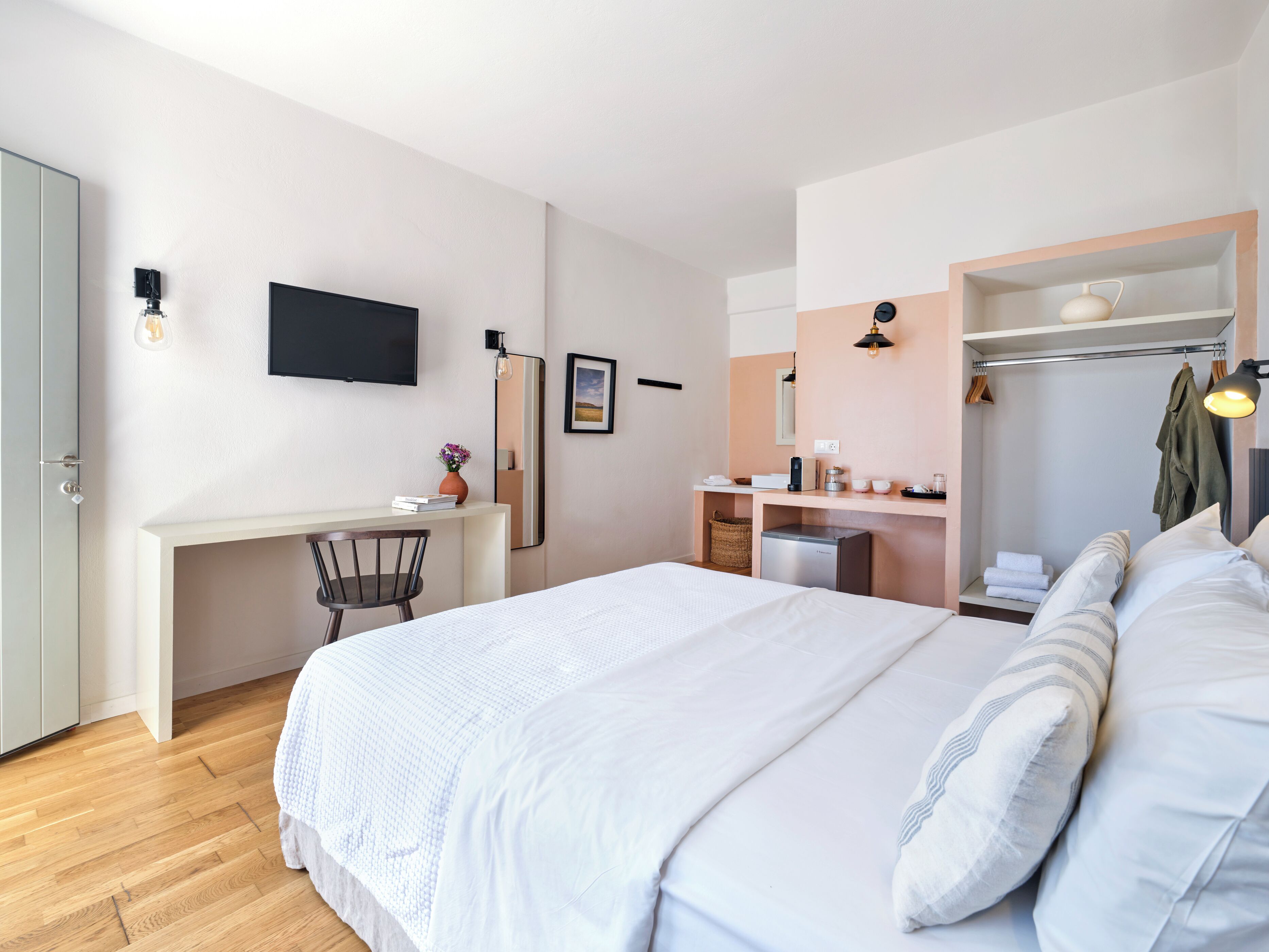 A modern bedroom with a large bed, a desk, a chair, a television, a mirror, and a small kitchenette.