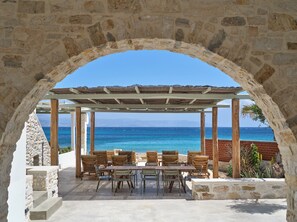 Un arc en pierre mène à un espace extérieur abrité, aménagé avec des tables et des chaises, d’où l’on peut admirer une mer d’un bleu limpide.