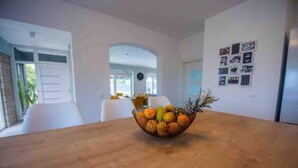 A dining area with a wooden table and a fruit bowl. There are white chairs around the table, and a white door is visible in the background. - Villa Vida (Zmijavci)