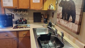 A kitchen with wooden cabinets, a coffee maker, a sink with a dish rack, and a wall-mounted painting of a bear.