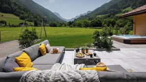 Un espace extérieur accueillant avec un canapé, des coussins et une couverture. On aperçoit un jacuzzi à l’arrière-plan, entouré d’un paysage verdoyant et de montagnes.