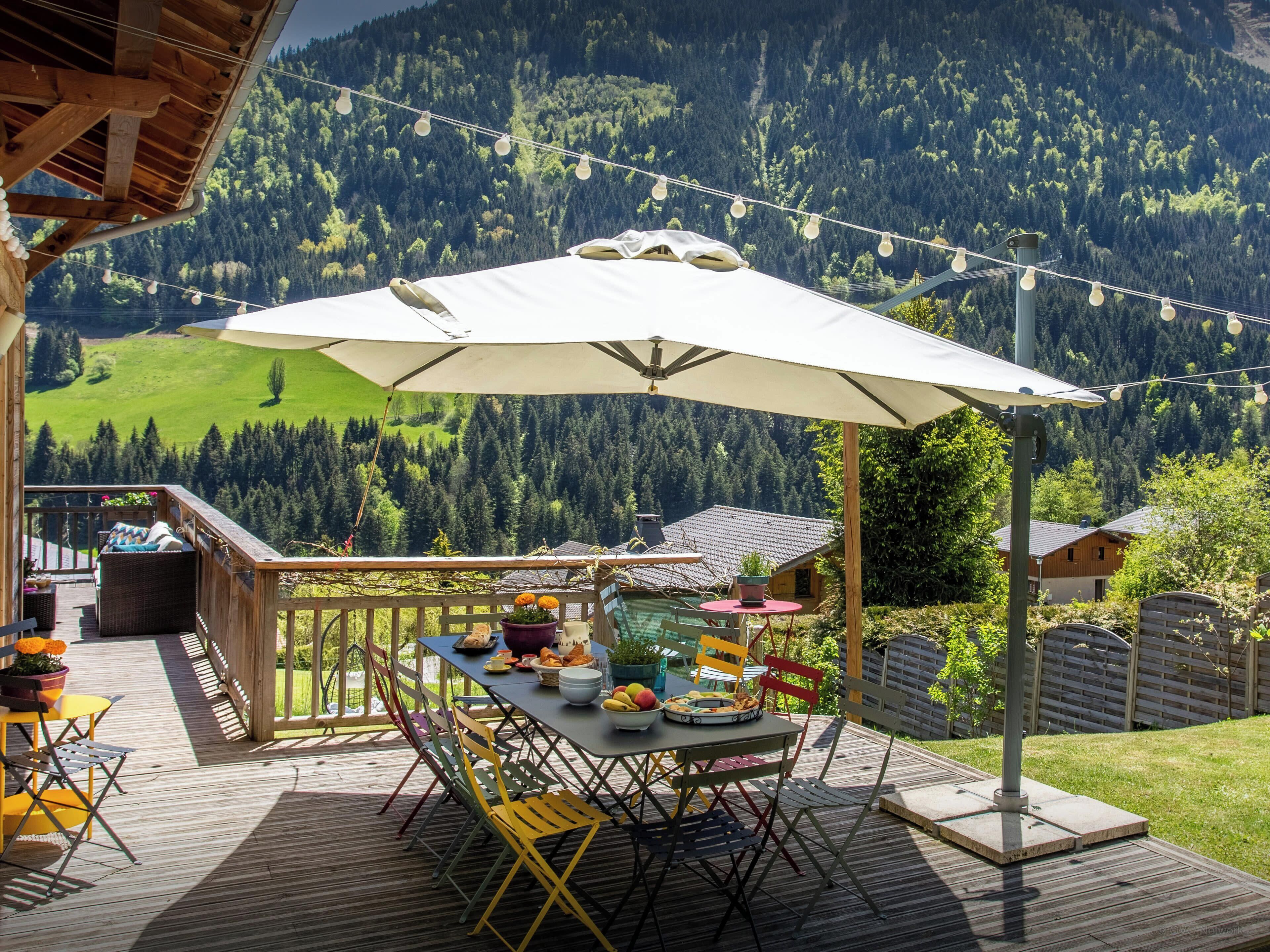 Een houten terras met een tafel, stoelen en een parasol, met uitzicht op een bebost gebergte.