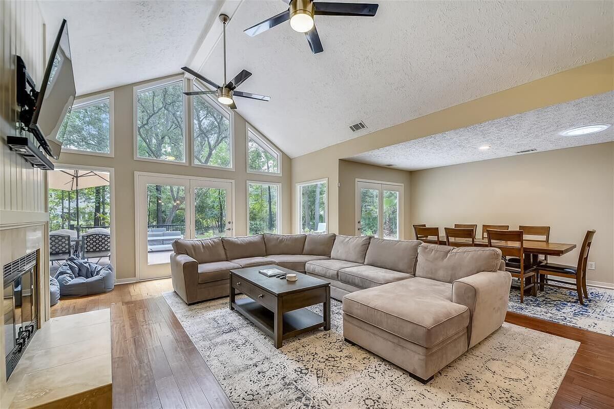 A spacious living room with a large sectional sofa, a coffee table, and a dining area with a table and chairs. There are large windows providing natural light and a view of the outdoors.