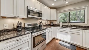 Une cuisine moderne avec des armoires blanches, un comptoir en granit, un four à micro-ondes encastré, une cuisinière et une fenêtre donnant sur les arbres.