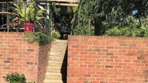 Una escalera de ladrillo que sube a una entrada con puerta, rodeada de follaje y una cerca de metal.