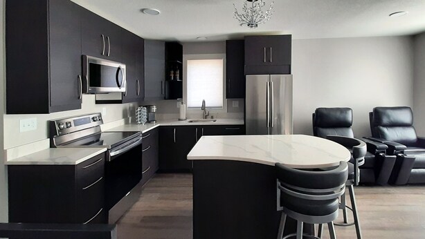 A modern kitchen with dark cabinetry, stainless steel appliances, a central island with bar stools, and a seating area with two black leather chairs. - Beautifully Designed Space. One Mile to Mayo Clinic. (Rochester)