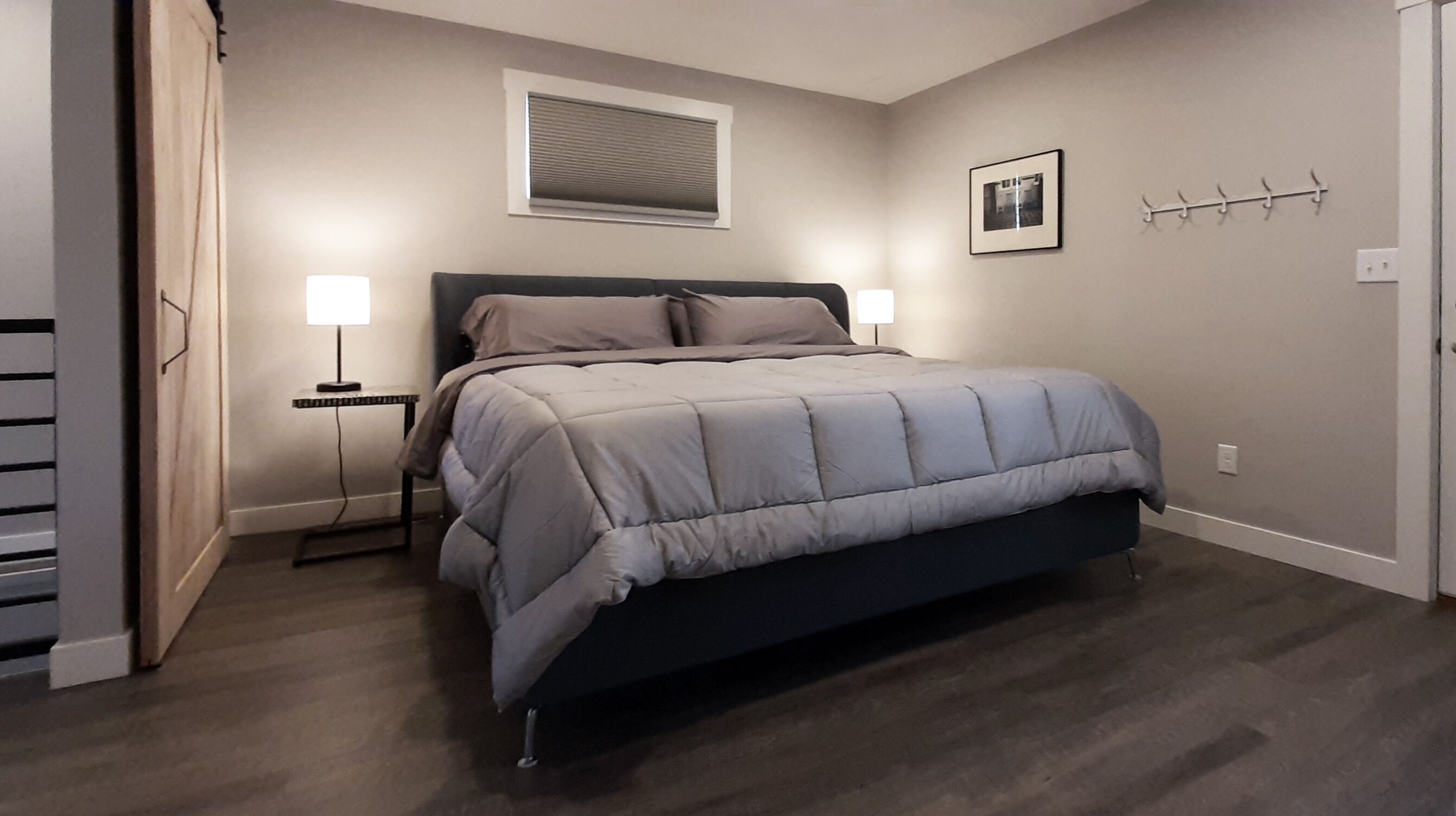 A bedroom with a bed, bedside lamps, a window with blinds, a framed picture on the wall, and a coat rack.
