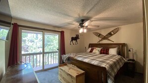 A bedroom with a bed, a ceiling fan, a wooden bench, and a large window with a view of greenery. - Hook and Antler (West Yellowstone)