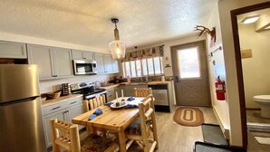 A compact kitchen with white cabinets, a stainless steel refrigerator, a microwave, and a wooden dining table with chairs. - Hook and Antler (West Yellowstone)
