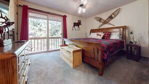A bedroom with a wooden bed, a dresser, a nightstand, and a large window with a view of trees. - Hook and Antler (West Yellowstone)